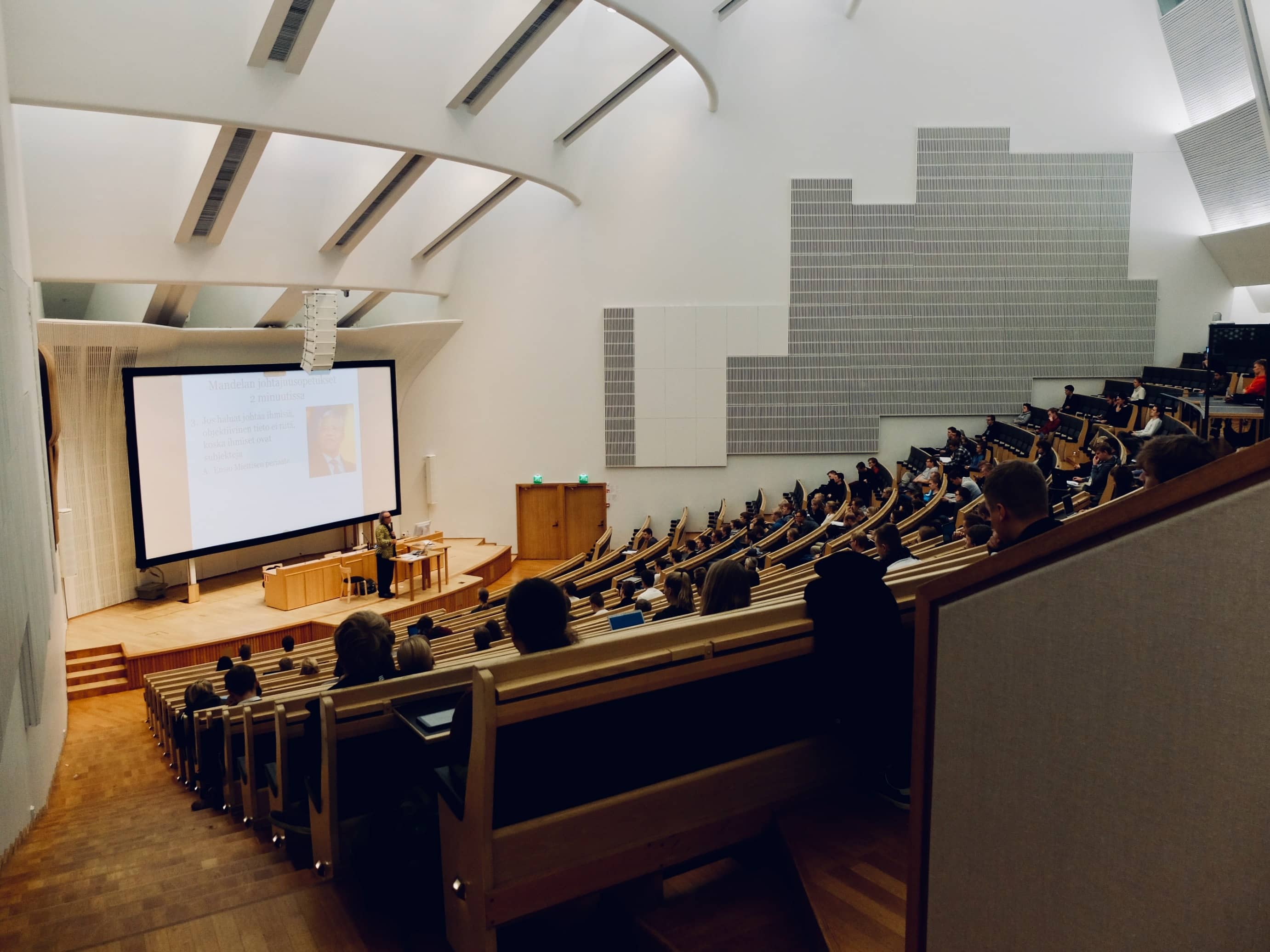 Ampitheatre style college classroom with students in chairs facing a lecturer on stage with projector behind.