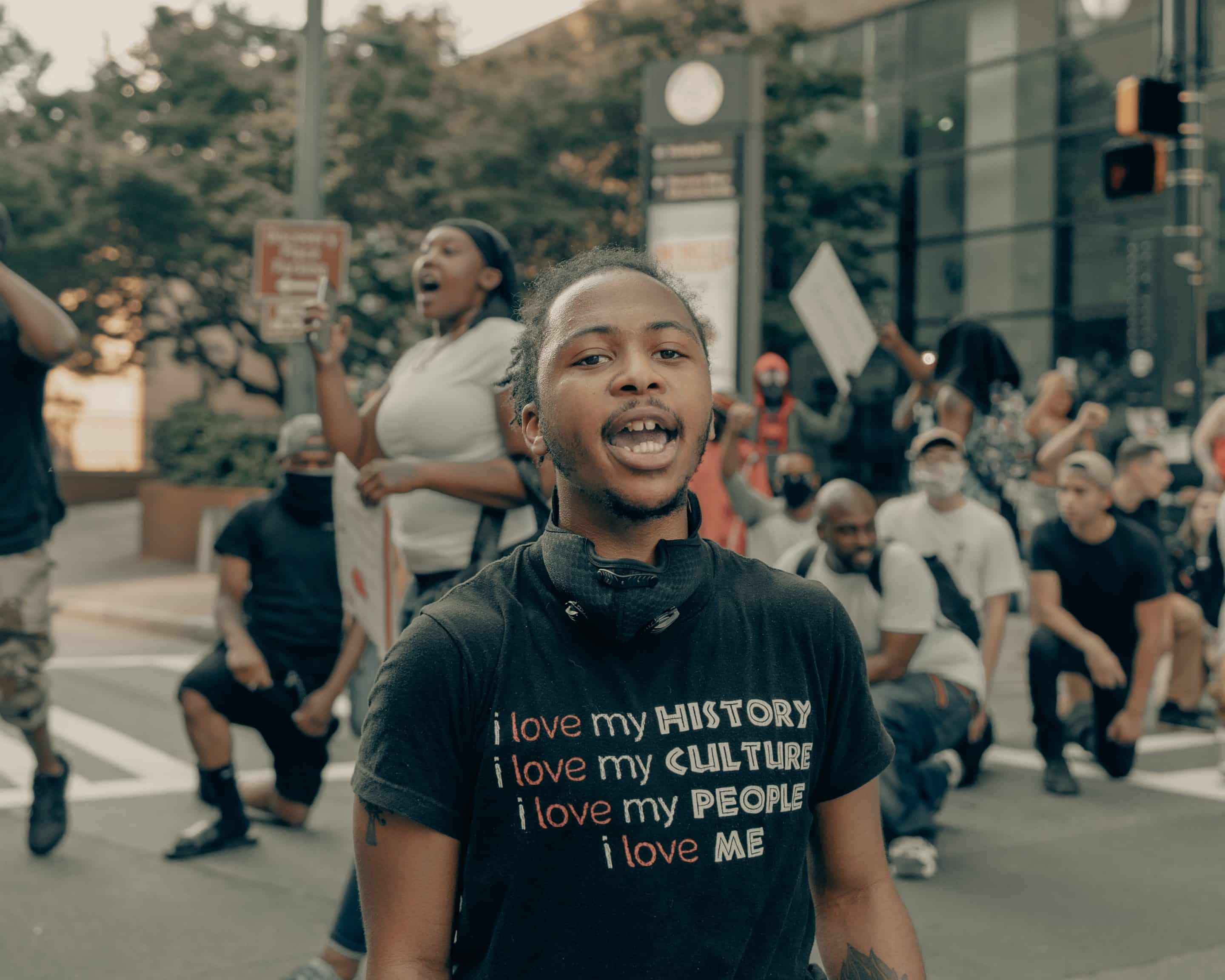 Young black man stands among crowd at George Floyd protests, wearing a black shirt that reads, "I love my history, I love my culture, I love my people, I love me"