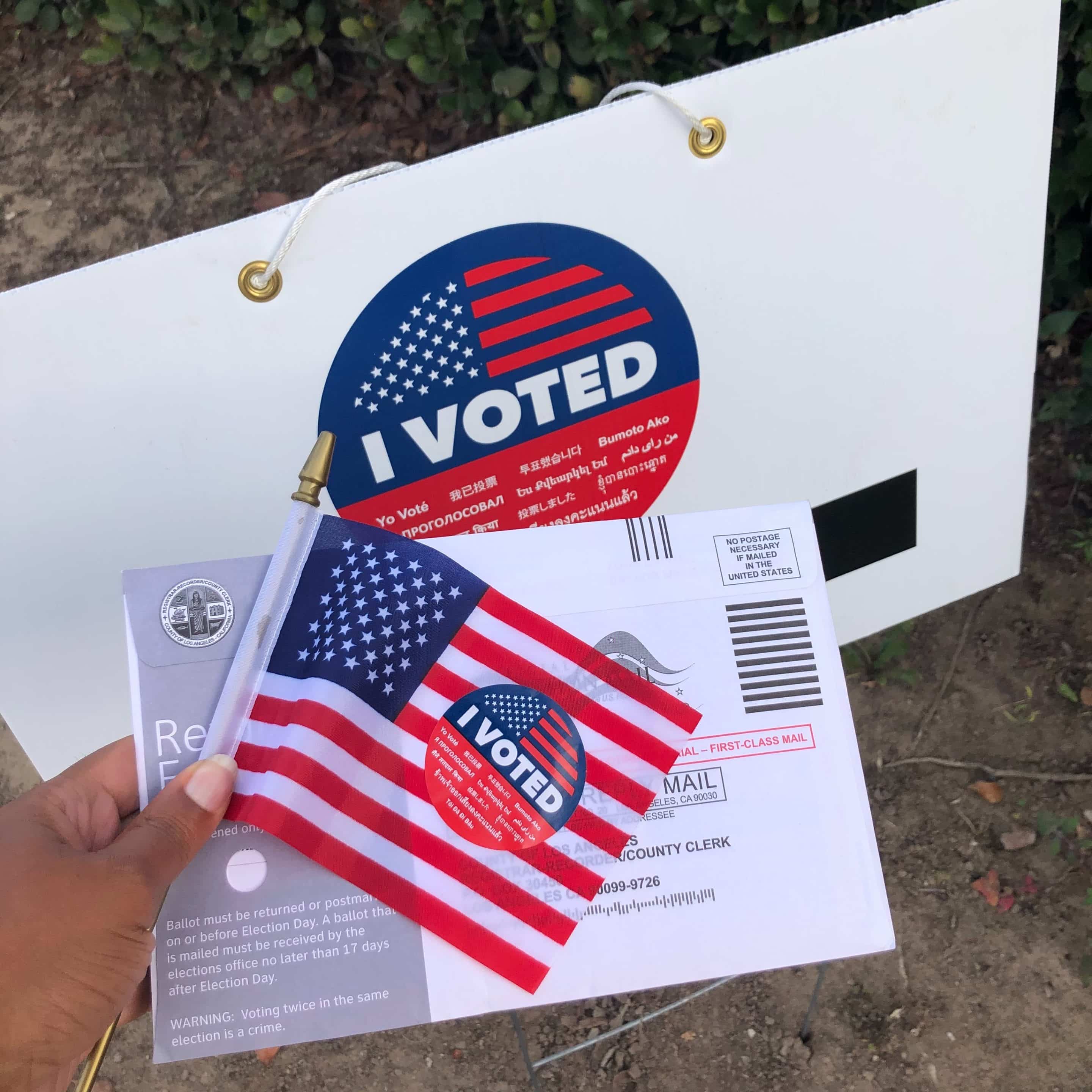 Black hand with pink nails holds an American flag, ballot, and 'I voted' sticker in front of a large 'I voted' sign