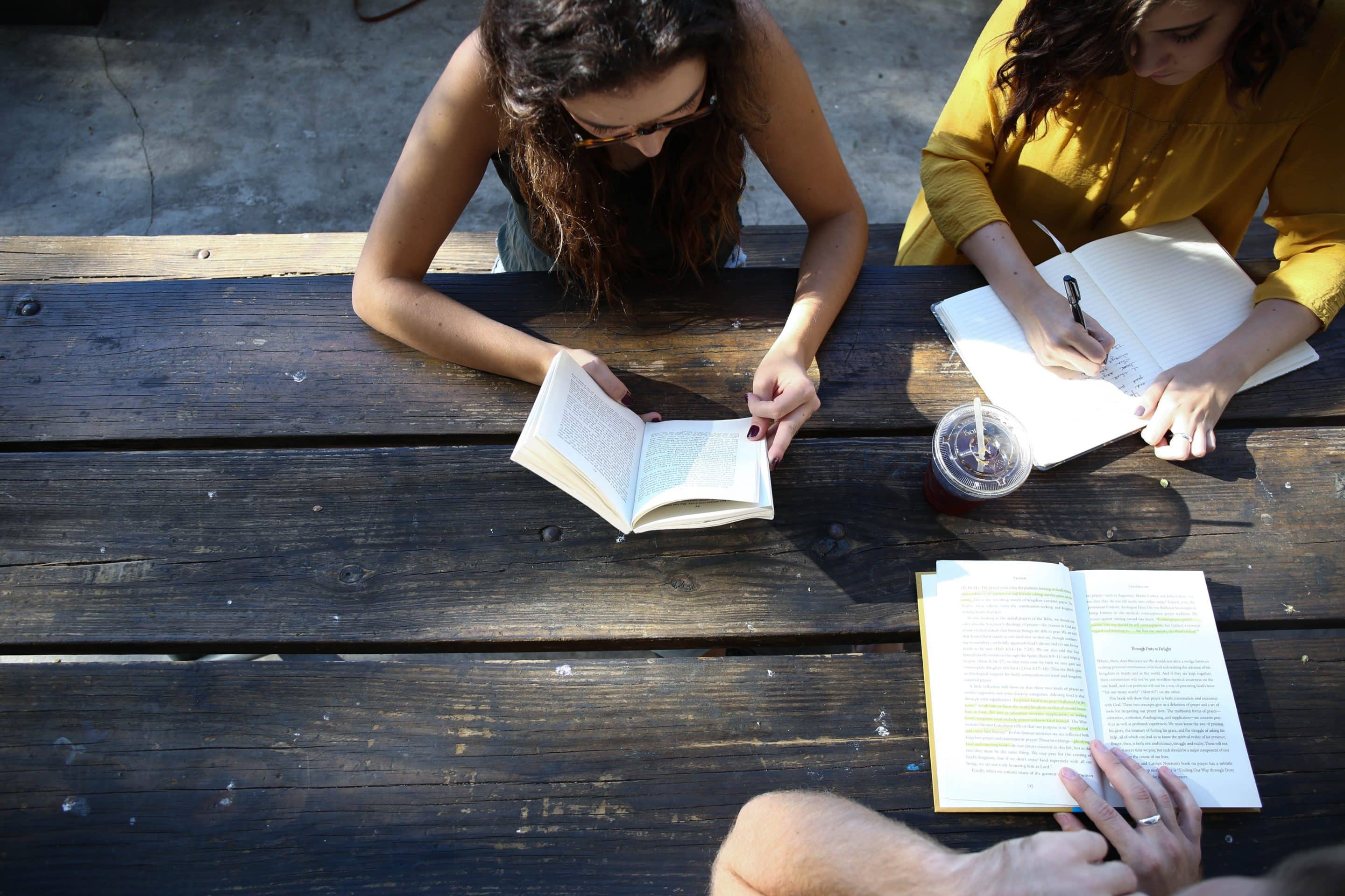 three women sit around a picnic table, discussing and writing in notebooks